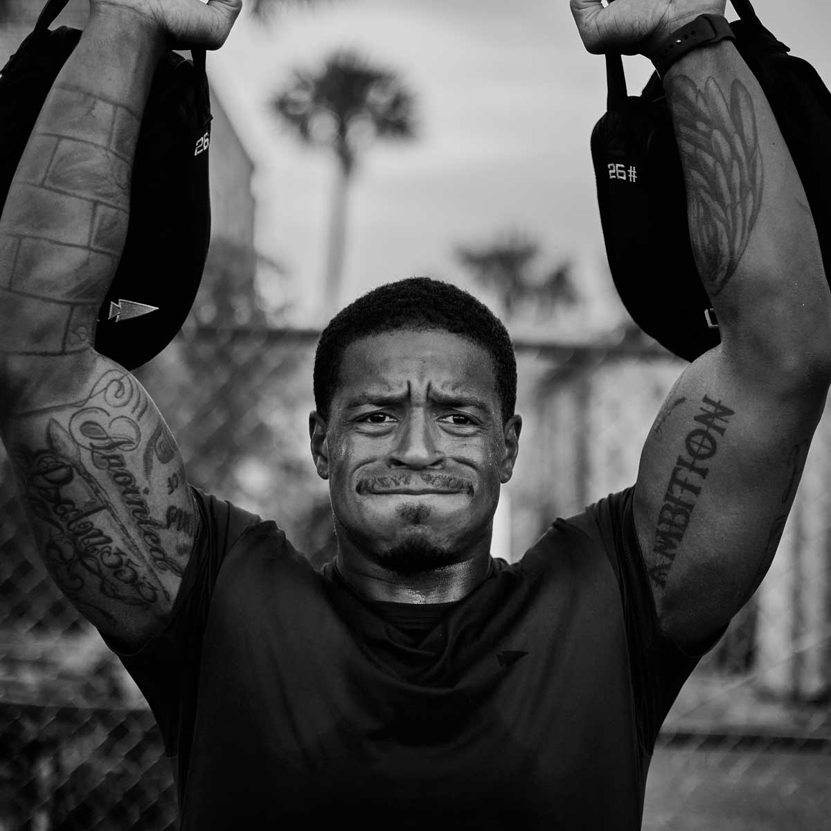 A man lifts weights overhead outdoors, showing determination and tattoos on his arms.