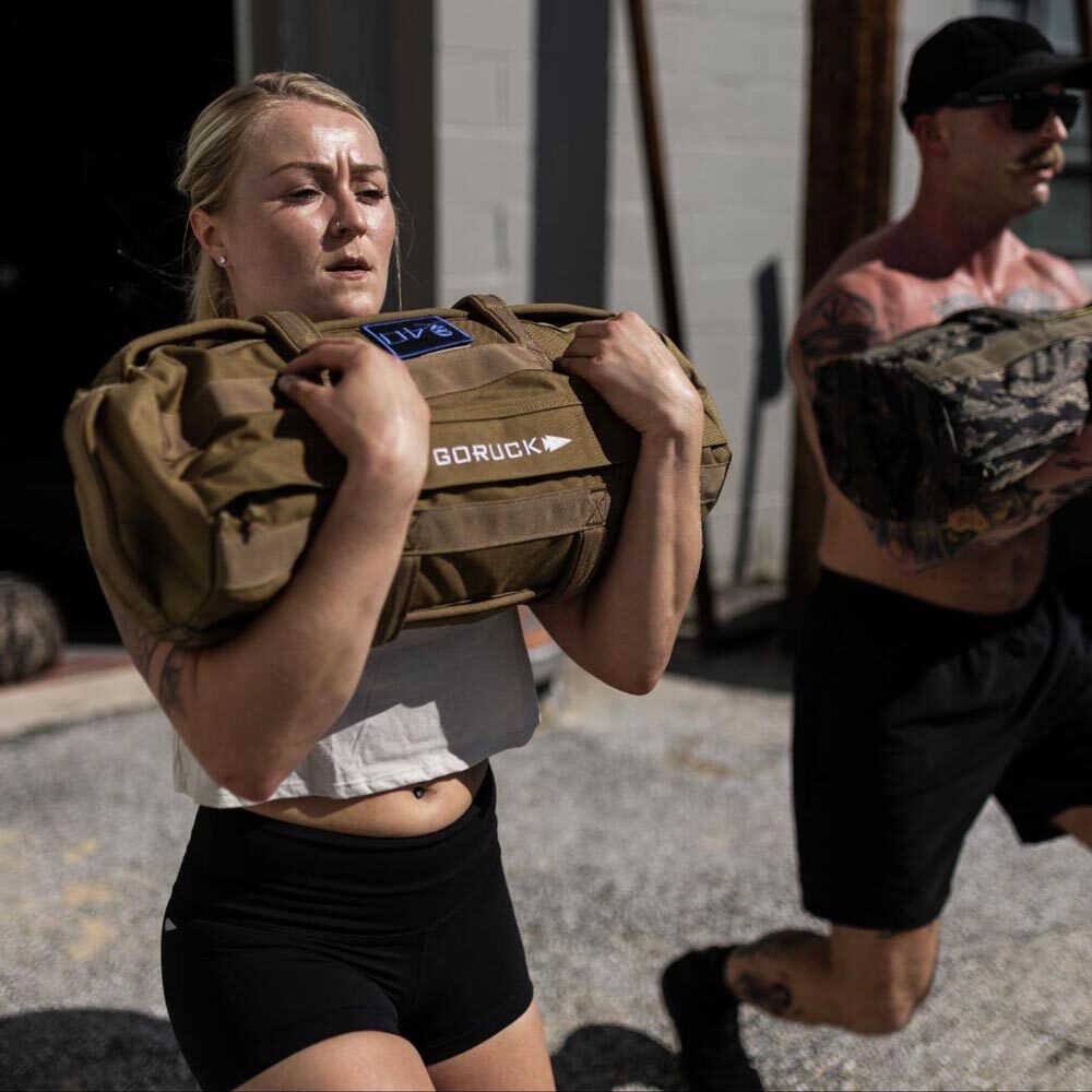 Two people exercise outdoors, holding weighted sandbags on their shoulders in sunlight.