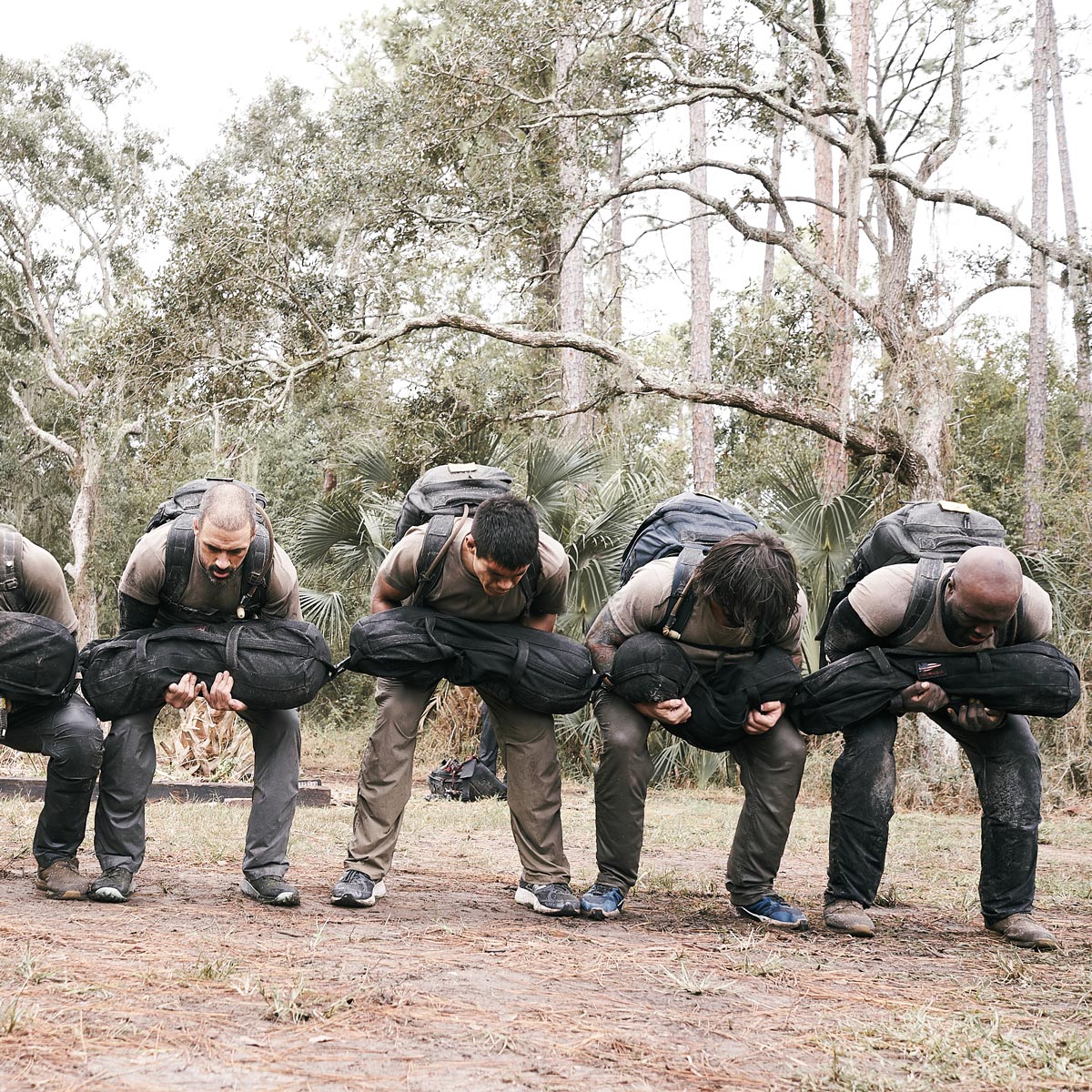 Five people outdoors bending over, holding heavy sandbags in front of them, surrounded by trees.