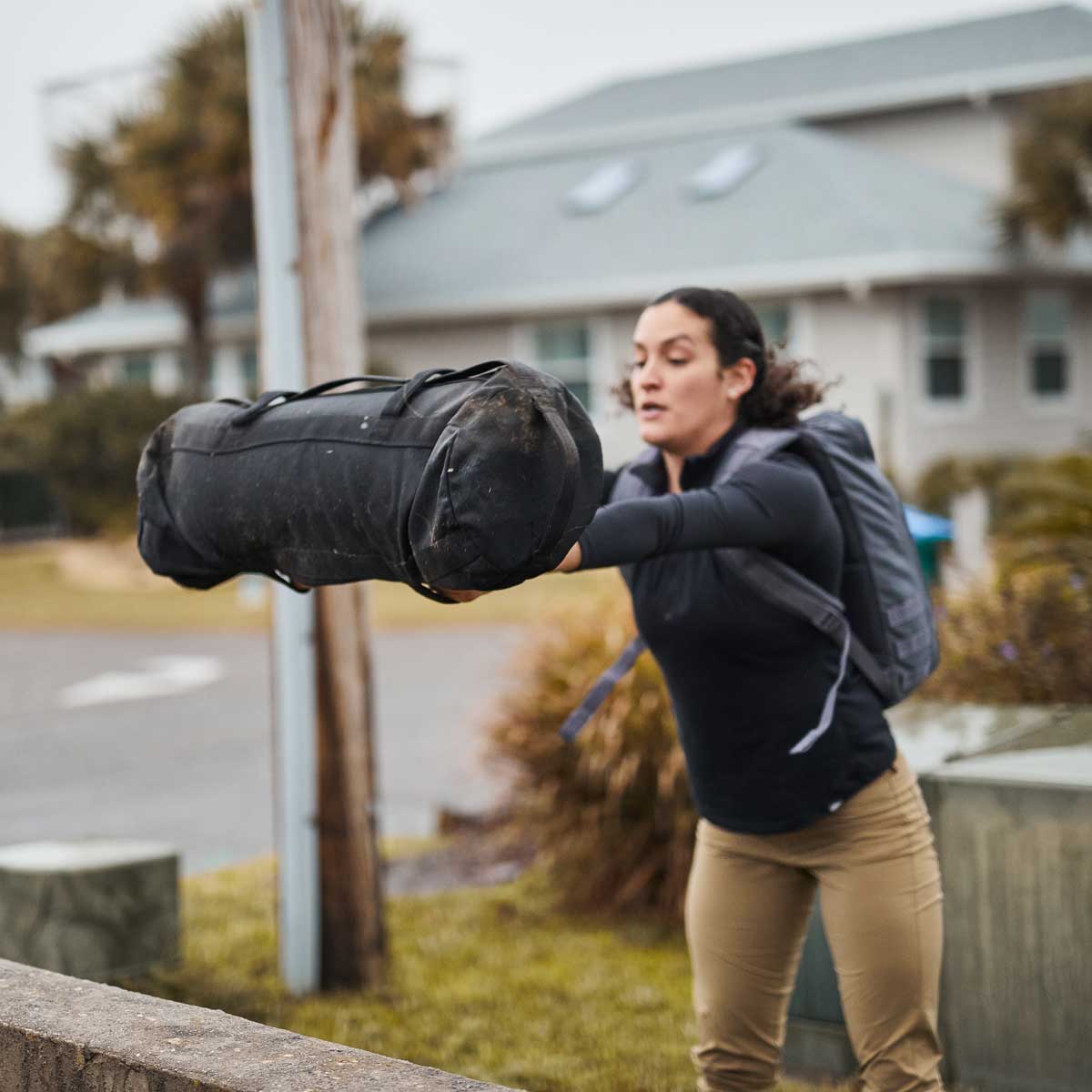Woman wearing a backpack throws a heavy sandbag outdoors near a house and utility pole.