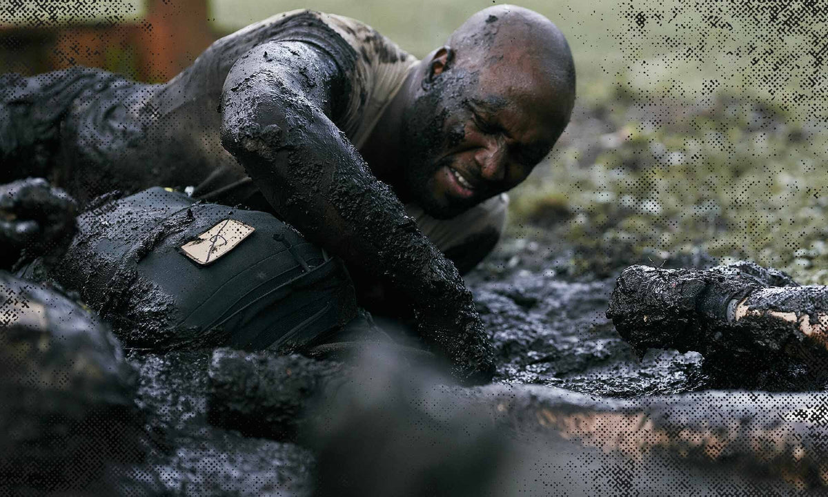 Man covered in mud struggles through an obstacle course, crawling on the ground outdoors.