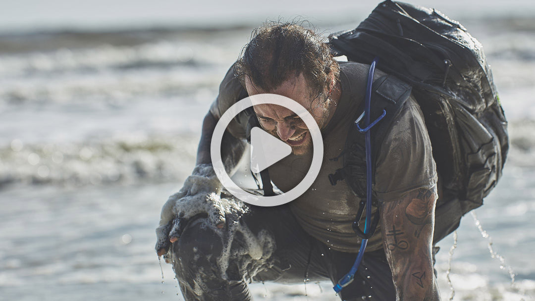 A person kneels on a beach, wet and dirty, carrying a large black backpack.