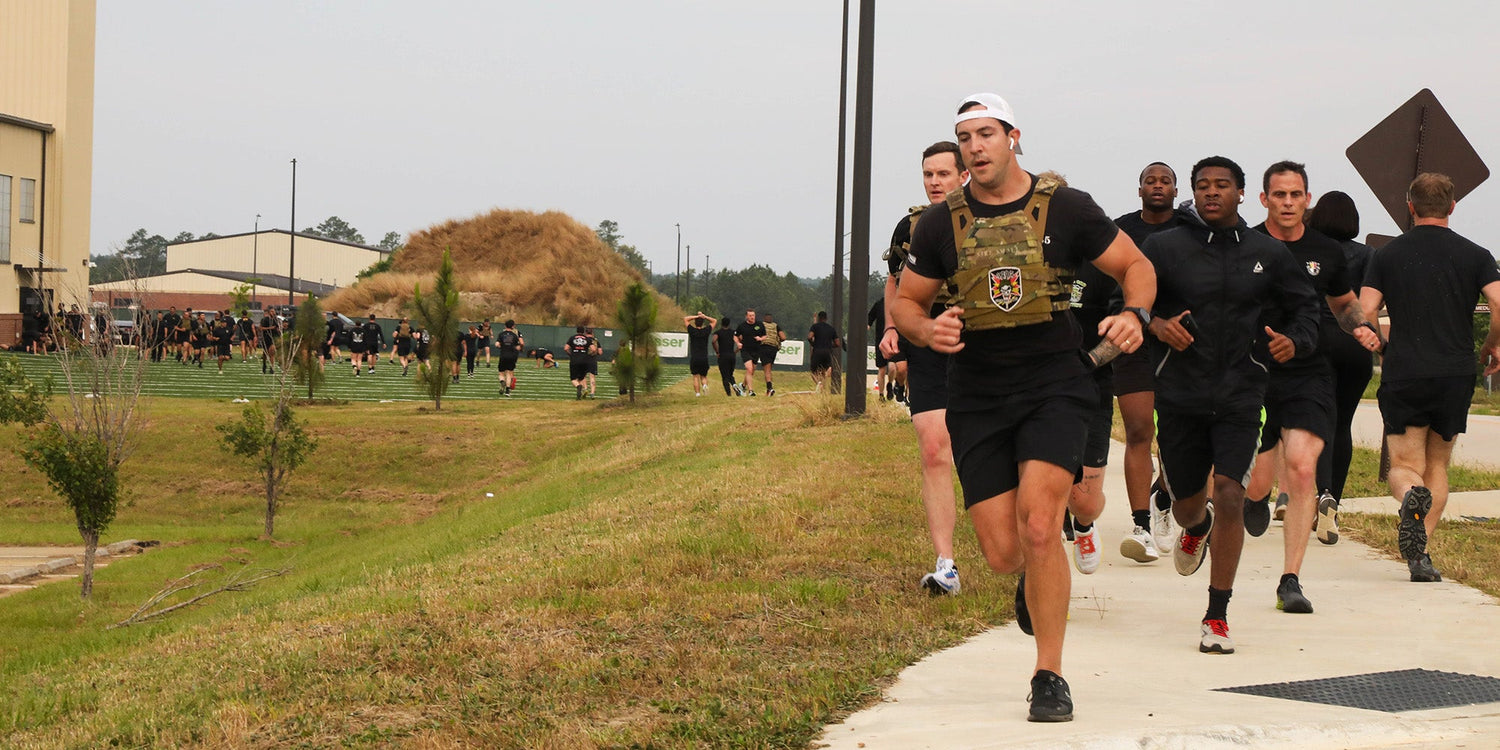 A group of people in athletic gear run together on a sidewalk near a grassy area and some buildings.