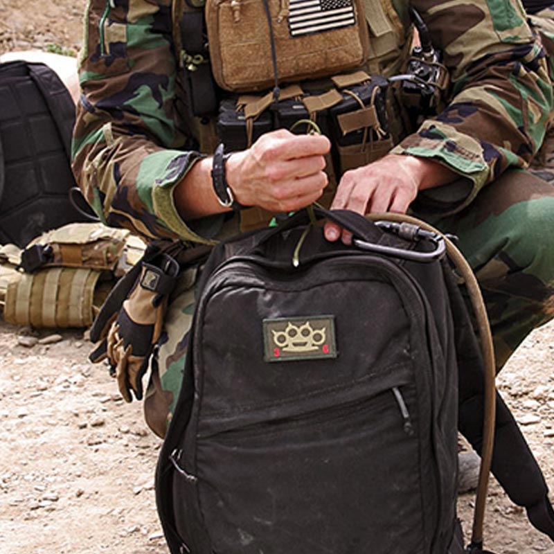 Person in camouflage holding a black backpack with a patch, kneeling on rocky ground with gear nearby.