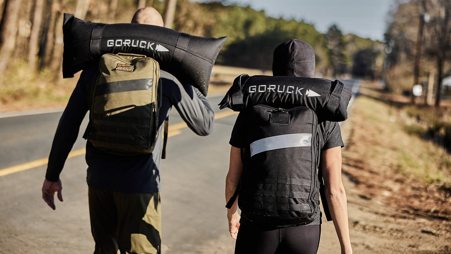 Two people walk on a road carrying sandbags labeled 