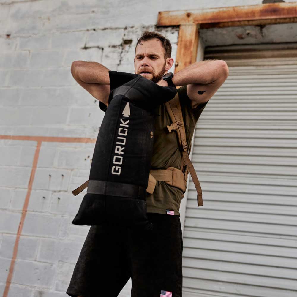 Man lifting a black GORUCK sandbag outside in front of a weathered building with a metal garage door.