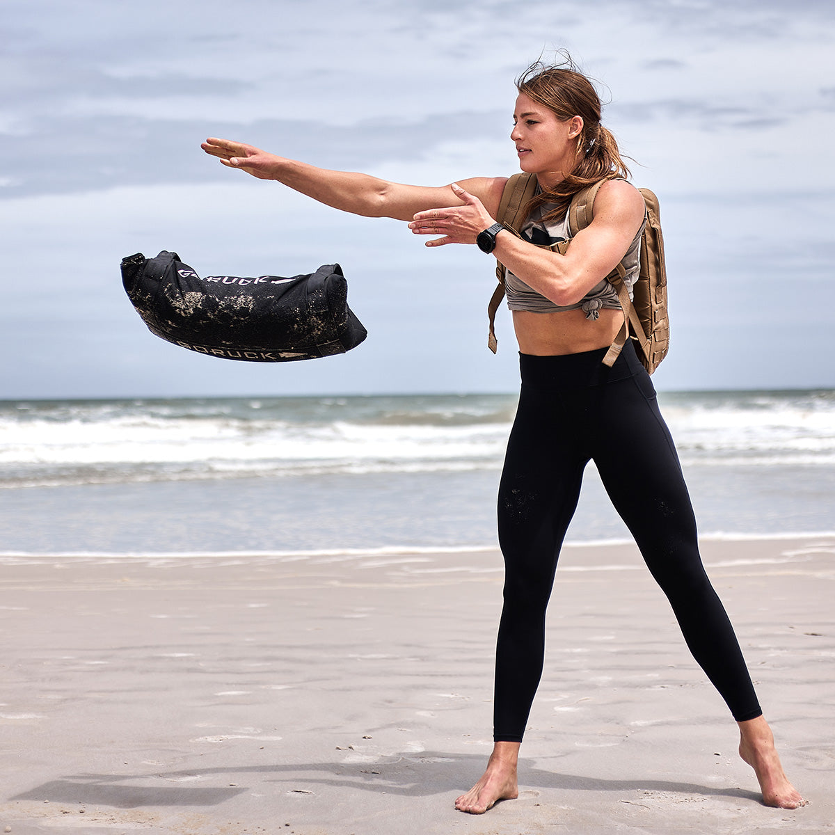 Woman throwing a sandbag on a beach, wearing workout clothes and a backpack, ocean in the background.