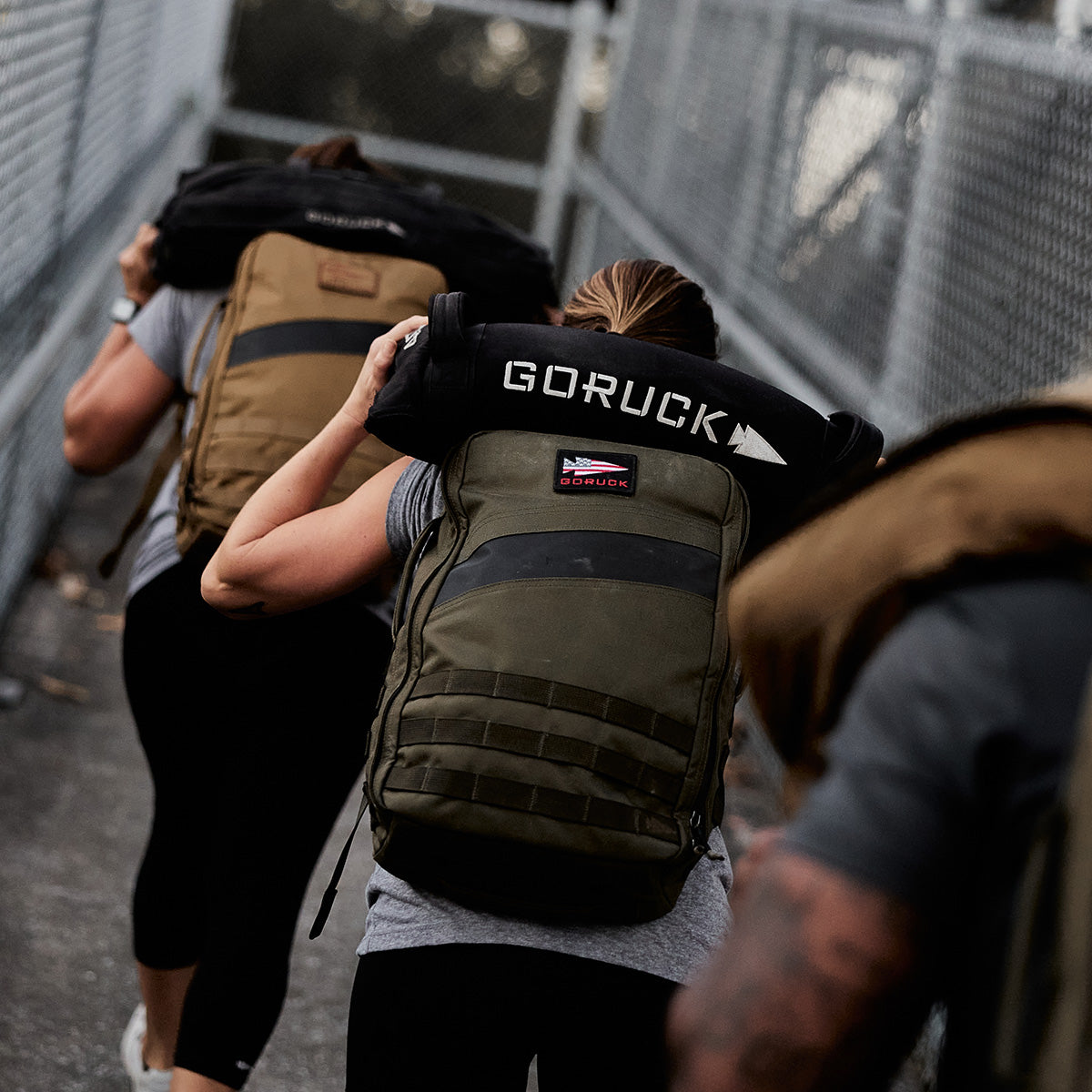 People outdoors carrying weighted rucksacks on their backs, participating in a GORUCK fitness event.