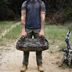 A person in a dark shirt and green Men’s Simple Pants by GORUCK stands on a dirt path enveloped by lush foliage, holding an abrasion-resistant, camouflage duffel bag. Tattoos are visible on their arms, with GORUCK's hiking gear made from Lightweight ToughDry® fabric nearby.