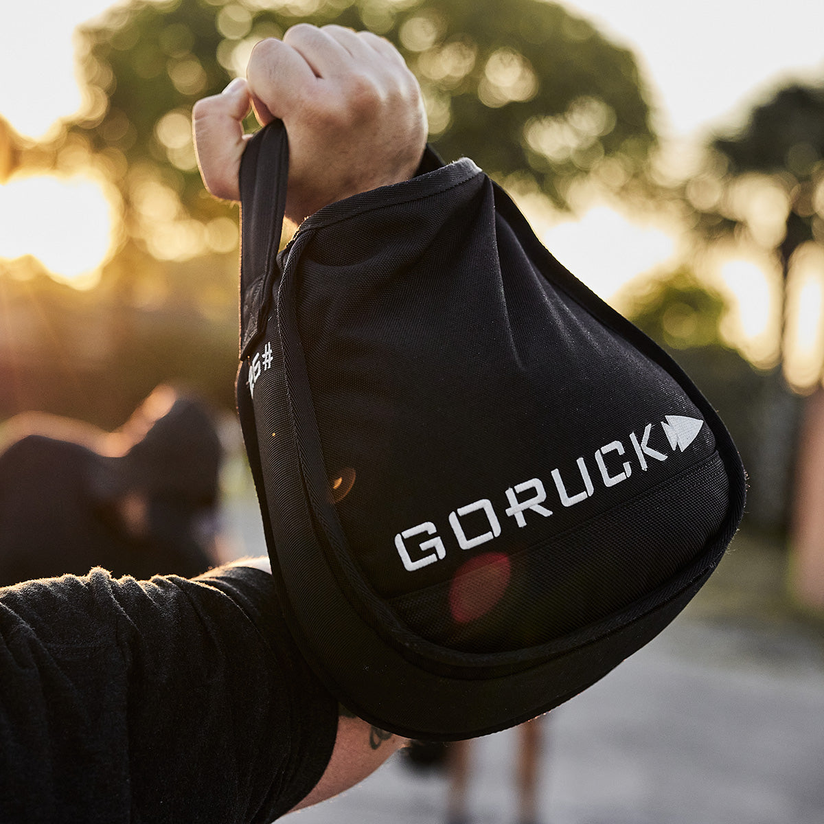 Person holding a black GORUCK kettlebell with trees and sunlight in the blurred background.