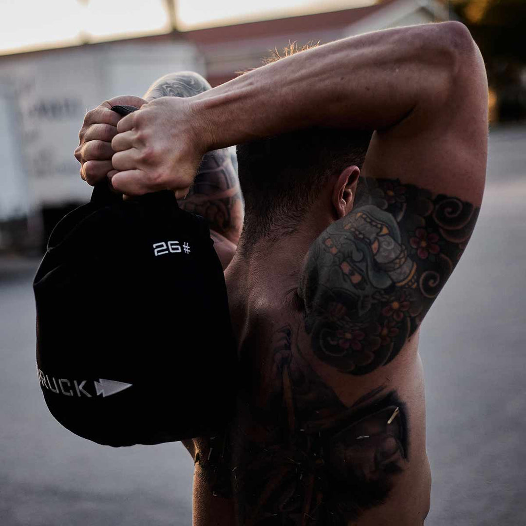 A man with arm tattoos skillfully lifts a 26-pound GORUCK Sand Kettlebell over his shoulders outdoors, highlighting its durable design.