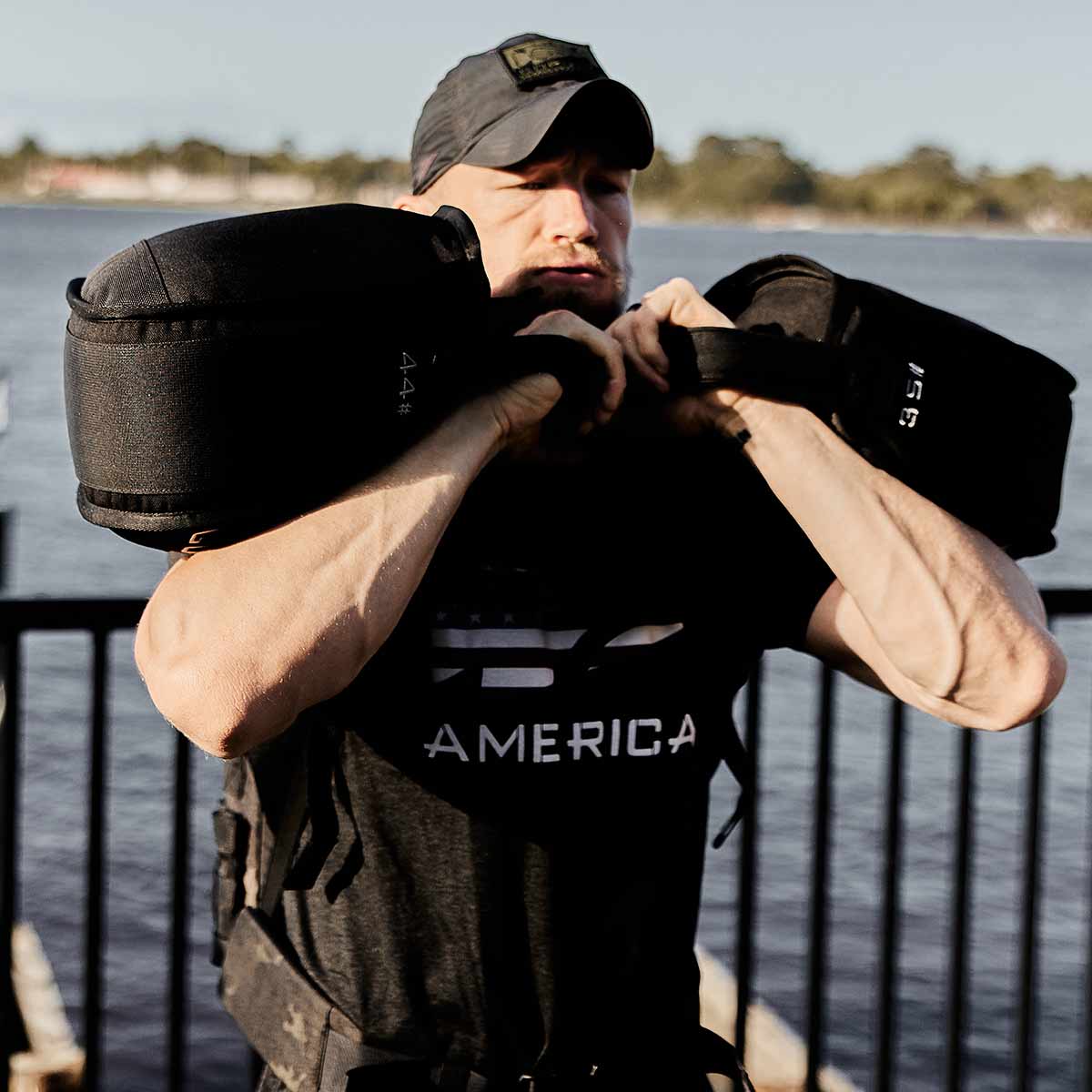 By the tranquil lake, an individual lifts a GORUCK Sand Kettlebell, pushing their endurance to new heights. Dressed in a black shirt with 