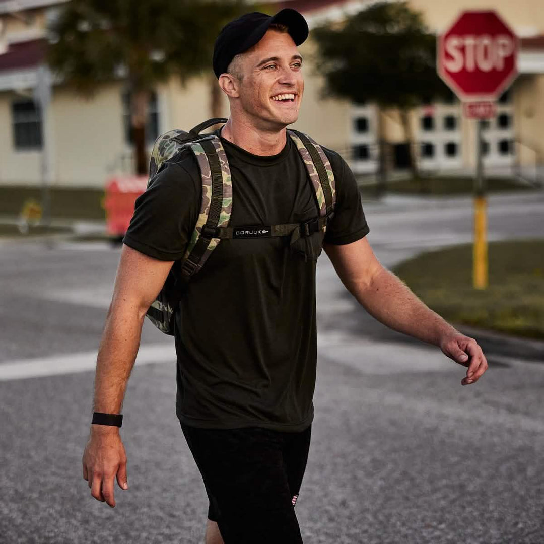 Smiling man rucking outdoors wearing GORUCK camo backpack and black t-shirt