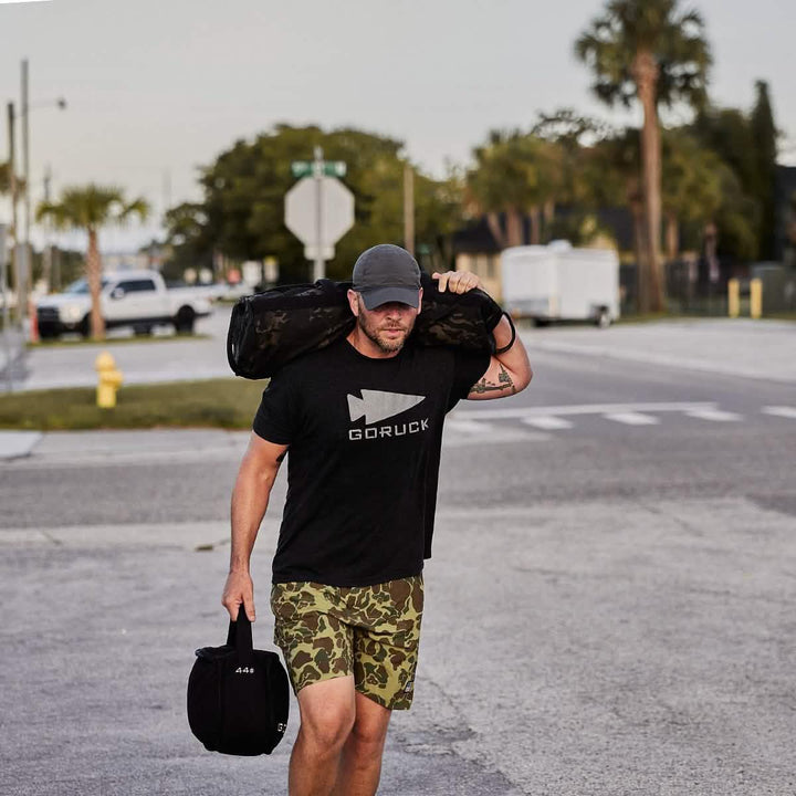 Man rucking outdoors wearing GORUCK shirt, camo shorts, carrying weighted gear bag.