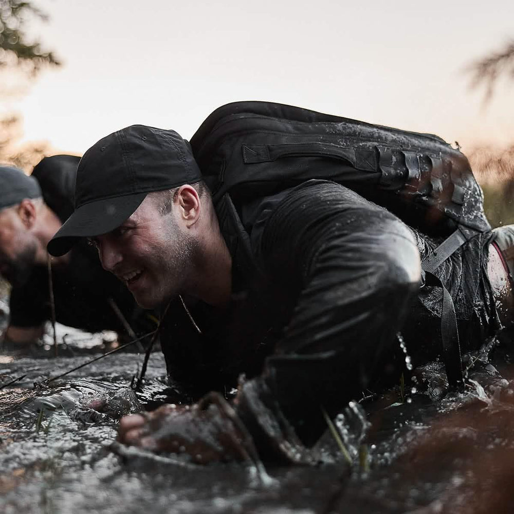 Man wearing GORUCK hat and backpack crawling through mud during intense ruck training