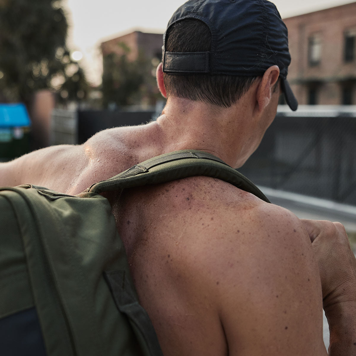 Shirtless man wearing a cap and backpack outdoors, with sweat and blemishes visible on his back and shoulders.