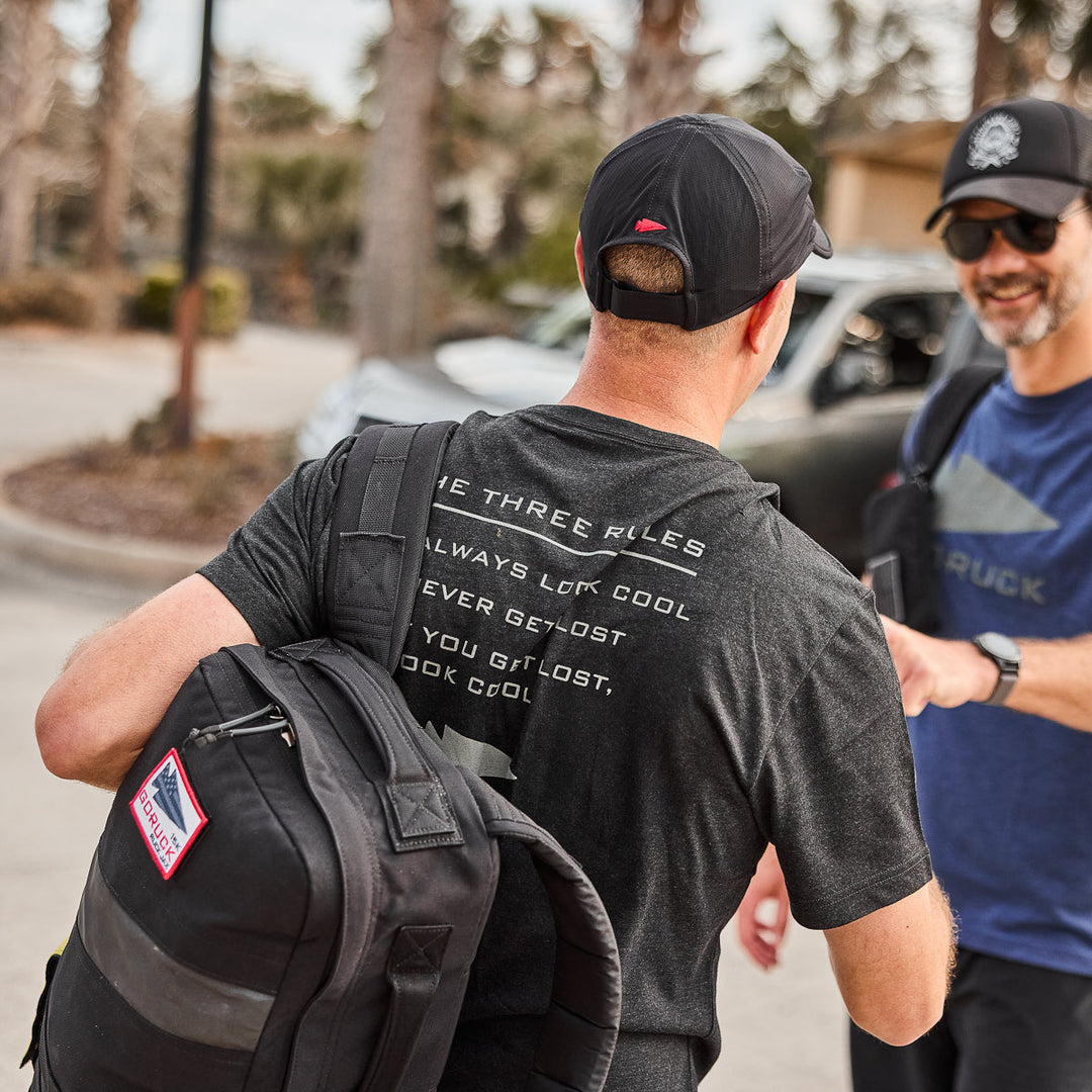 Two men in the vendor-unknown Three Rules Tee - Tri-Blend and backpacks walk outdoors, smiling and talking on a sunny day—captured in stunning photos.