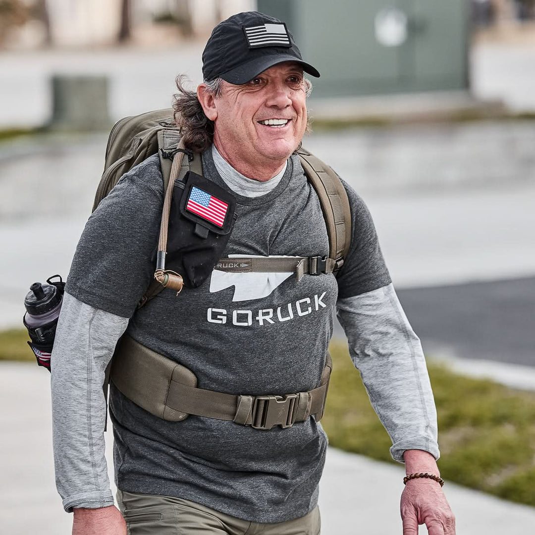 Smiling man outdoors wearing the vendor-unknown Three Rules Tee - Tri-Blend, backpack, and cap with an American flag patch—ideal for adventure photos.