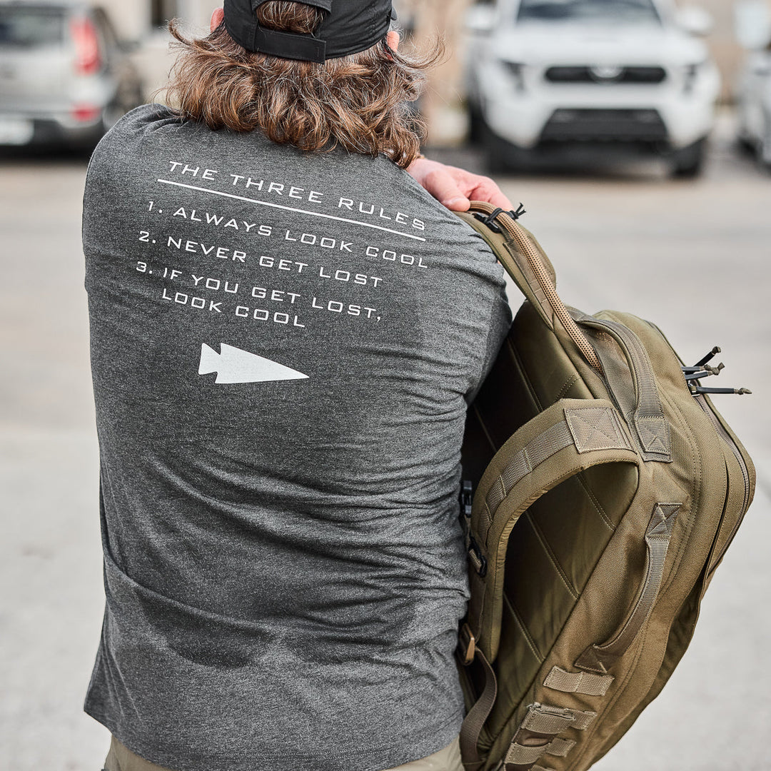 A man with long hair, cap, and backpack wears the vendor-unknown Three Rules Tee - Tri-Blend featuring "THE THREE RULES: Always look cool..."—as seen in semi-famous photos.