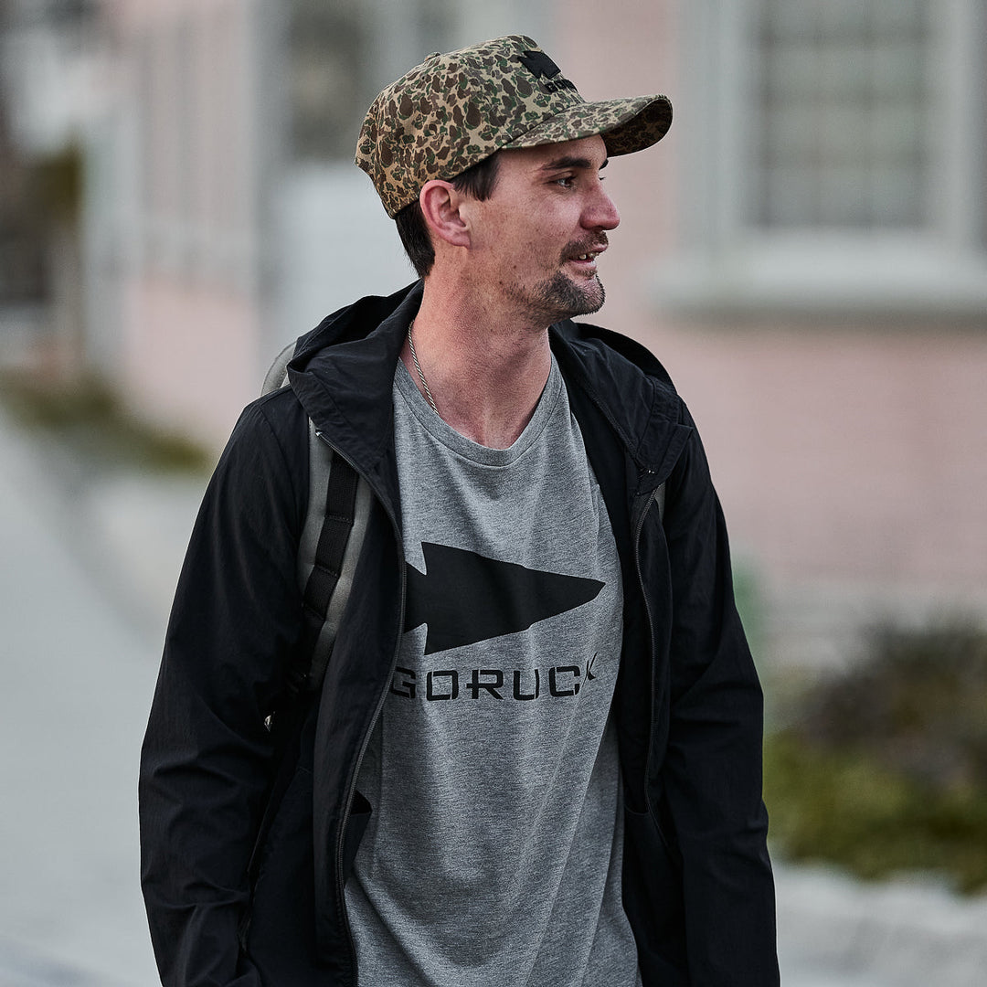 A man in a camo cap and black jacket walks outside, wearing the vendor-unknown Three Rules Tee - Tri-Blend in gray and carrying a backpack.