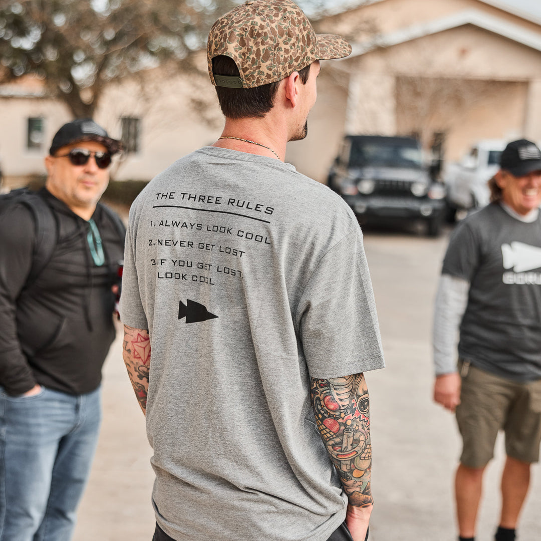 A man wearing the vendor-unknown Three Rules Tee - Tri-Blend, featuring "The Three Rules" printed on the back, poses for photos with friends outdoors.