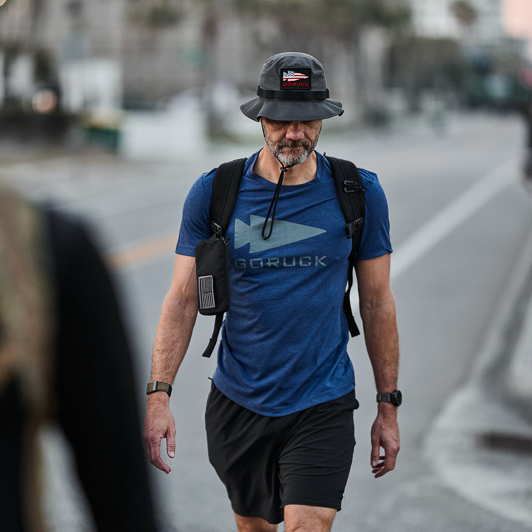 A man in the vendor-unknown Three Rules Tee - Tri-Blend (blue) and a black hat walks outside with a backpack and water tube along a city street—an ideal look for candid photos.