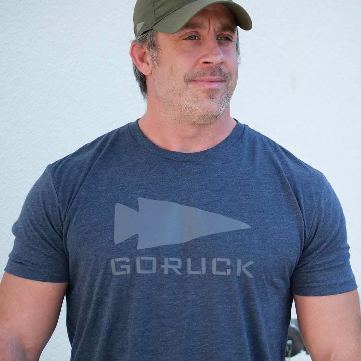 A semi-famous man wears a vendor-unknown Three Rules Tee - Tri-Blend in blue and a green cap, standing before a light wall, ready for photos.