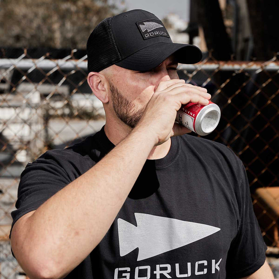 A man in a Performance Trucker Hat - GORUCK Spearhead and a GORUCK t-shirt, sipping from a can by a chain-link fence, showcasing his style with an adjustable Velcro strap for comfort.