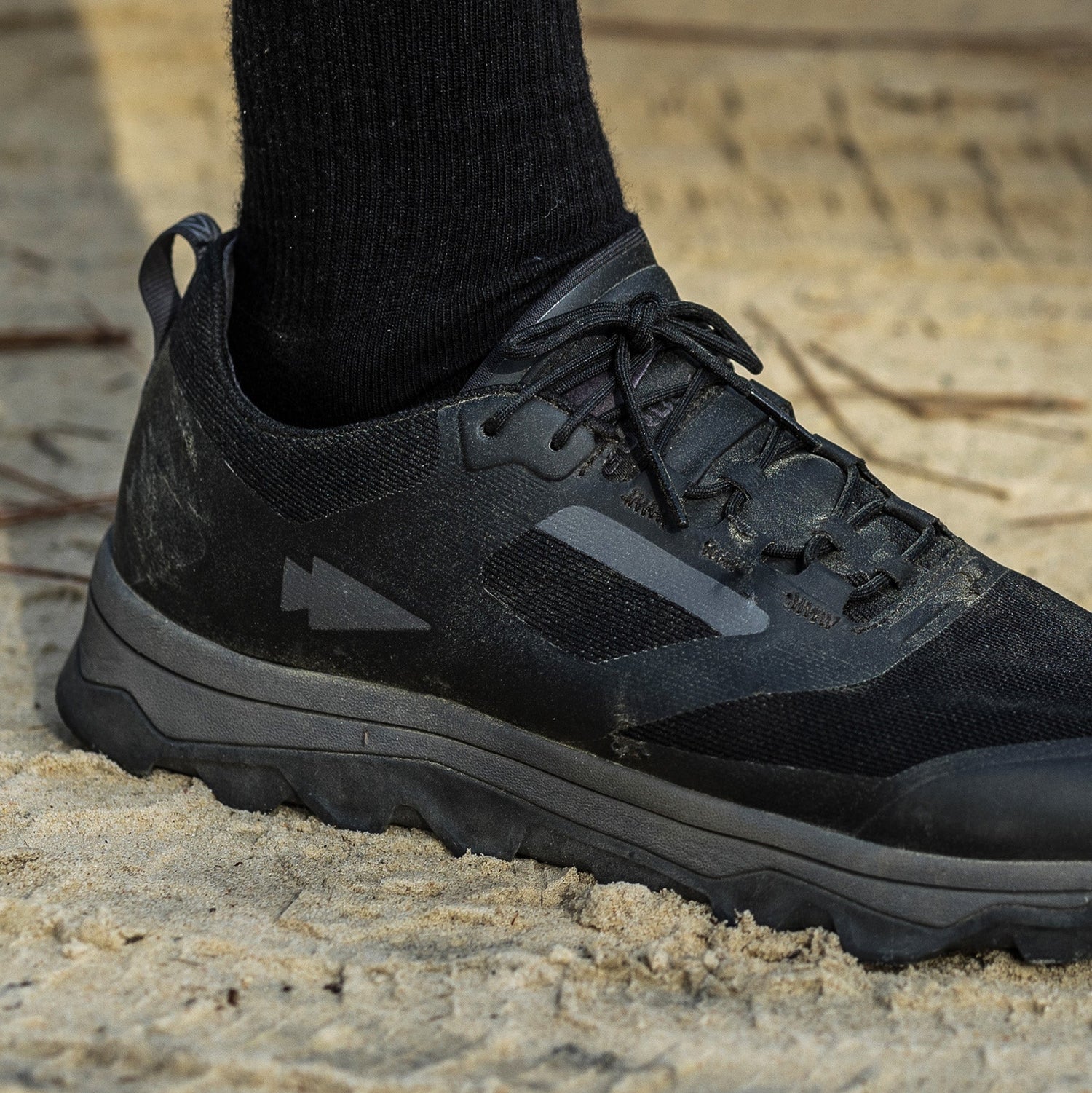 A person wearing a black sneaker and black sock stands on sandy ground.