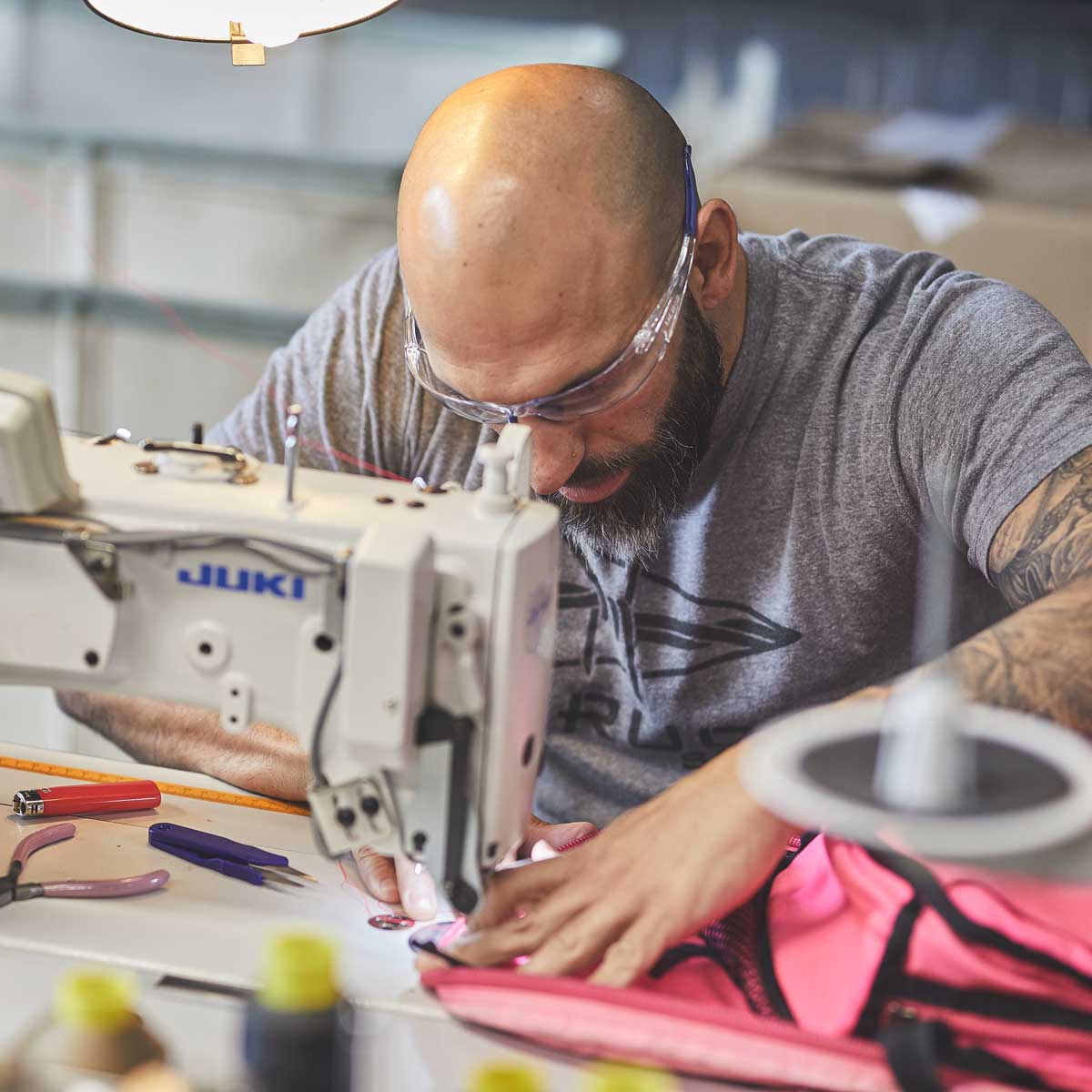 Bald man wearing goggles sewing pink fabric with an industrial sewing machine in a workshop.