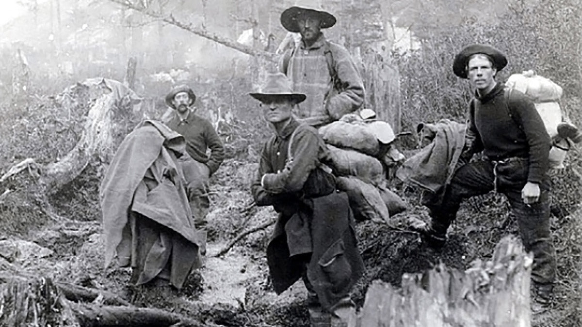 Four men in rugged outdoor clothing hike through a muddy, forested trail carrying heavy packs.