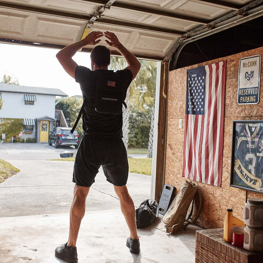 Man exercising in a garage with a backpack on, near an American flag and sports memorabilia on the wall.