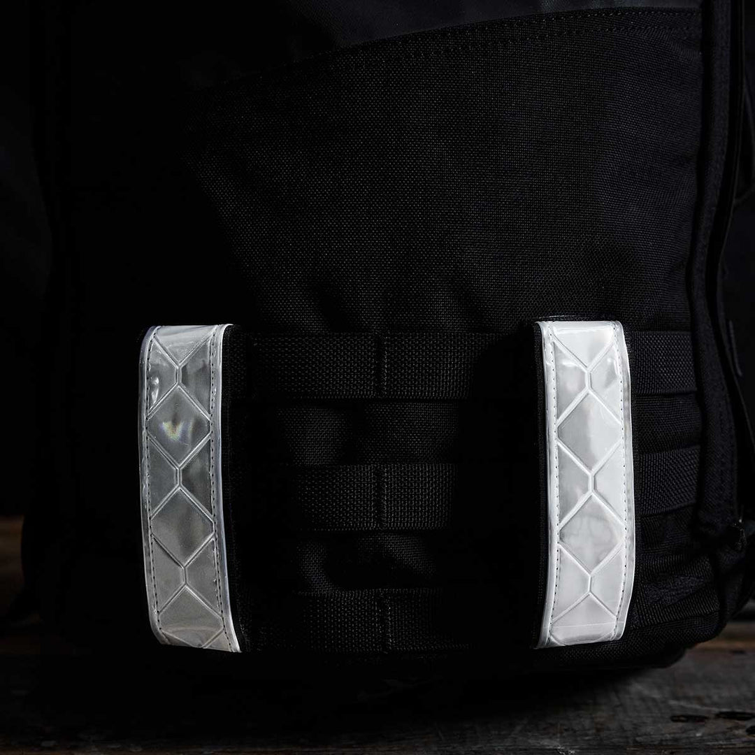 Close-up of the GORUCK Reflective Ruck Bands, a black backpack featuring MOLLE webbing and reflective straps, which enhance nighttime visibility against a dark background.