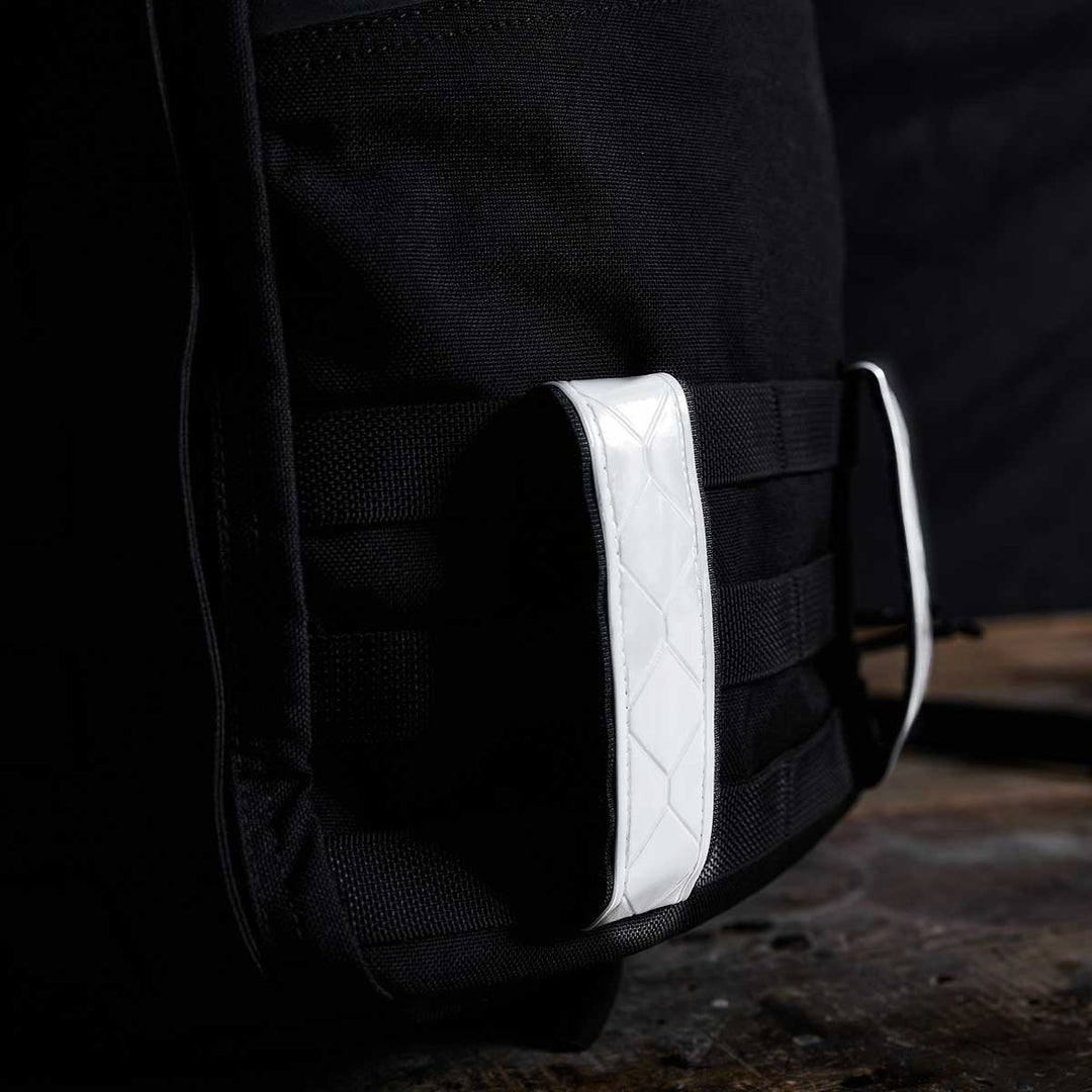 Close-up of a black backpack by GORUCK, showcasing Reflective Ruck Bands for improved visibility at night, complemented by a white, textured handle set against a dark background.