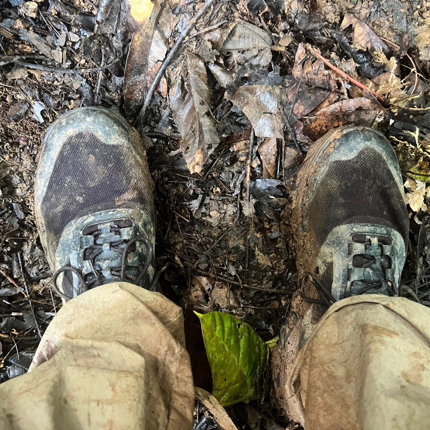 Muddy hiking boots and tan pants standing on a forest floor covered with leaves and dirt.