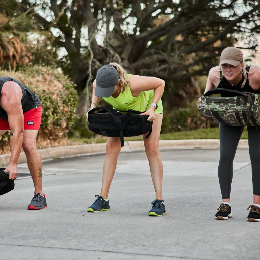 Three people outdoors, bending forward while lifting sandbags in a group workout, all wearing GORUCK Ballistic Trainers - Wolf Grey + Acid Lime W / Acid Lime Reflective Spearhead for intense routines.