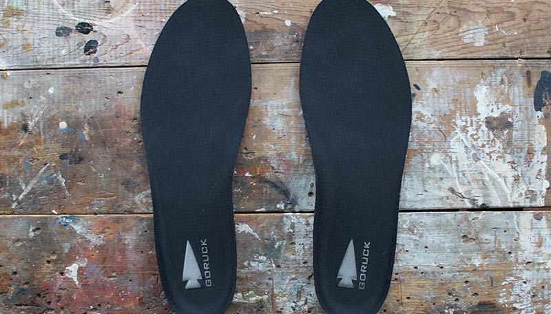 A pair of black GORUCK insoles on a paint-splattered wooden surface.