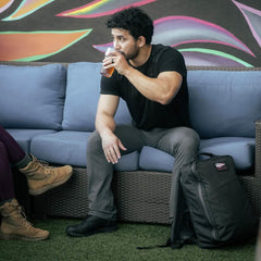 A man sits on a couch, sipping a drink with his waterproof GORUCK GR1 USA - X-PAC backpack featuring AquaGuard zippers beside him, set against a vibrant mural backdrop.