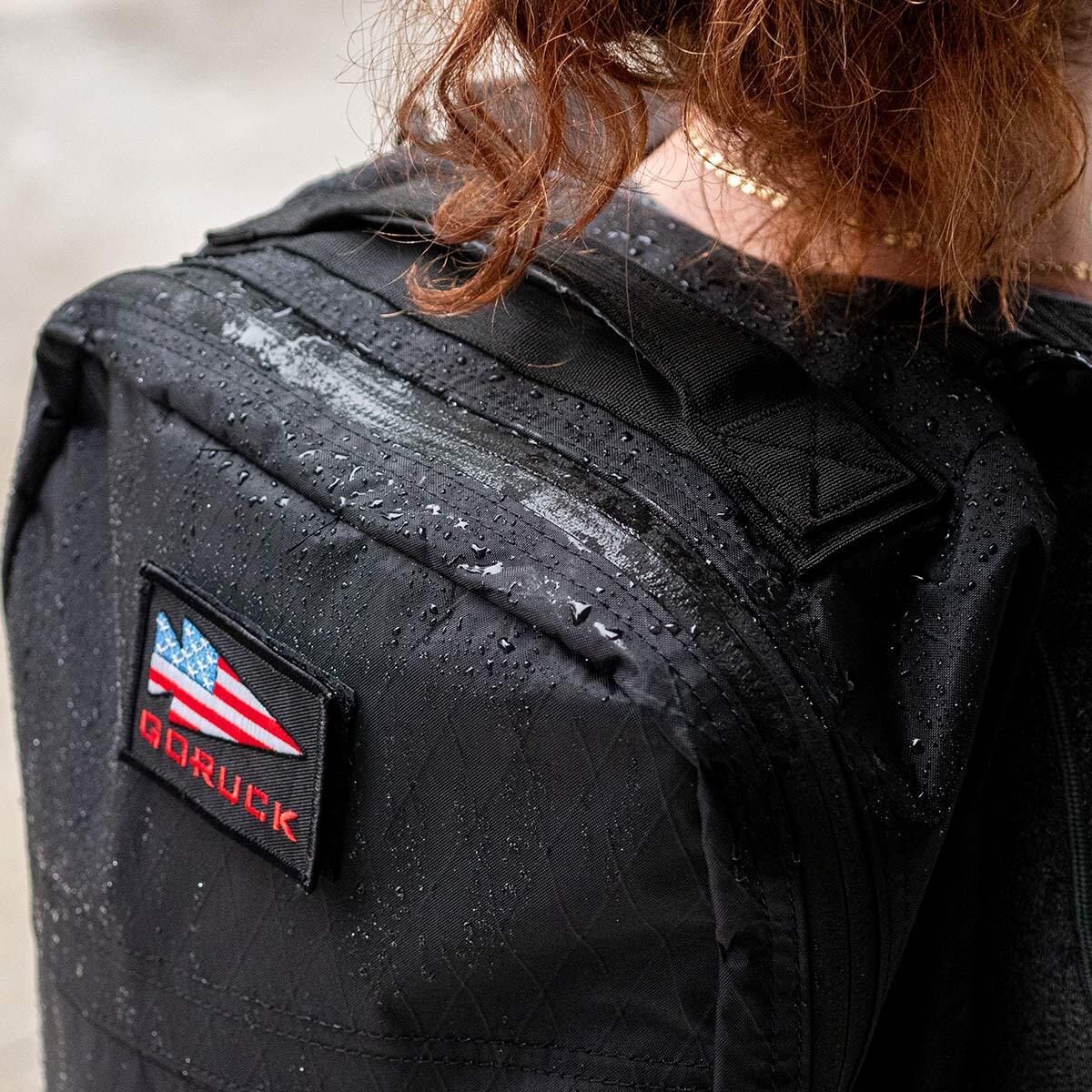 Close-up of a person wearing a black backpack with a GORUCK patch, wet from rain.